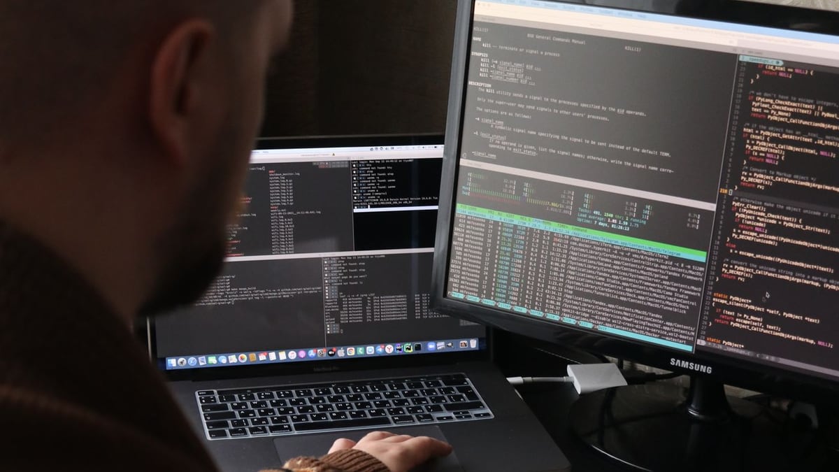 Developer at a desk working across a laptop and monitor filled with terminal windows and code